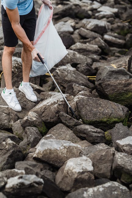 A person wearing white sneakers, black shorts, and a blue shirt is standing on a rocky shoreline, using a grabber tool to collect small pieces of litter from the uneven surface comprised of large, weathered stones with rough textures and various shades of gray, brown, and mossy green. The individual holds a white plastic bag in one hand, ready to deposit the collected waste, which suggests an activity related to cleanup efforts or waste collection in a natural outdoor environment. The background features more rocks and natural terrain, with no other objects or structures visible, emphasizing the focus on manual rubbish removal in a rugged outdoor setting. This scene, associated with private waste handling or outdoor rubbish clearance, highlights the importance of local community efforts and independent collection methods, aligning with services like those offered by House Clearance Bayswater in managing waste removal outside of standard municipal services.