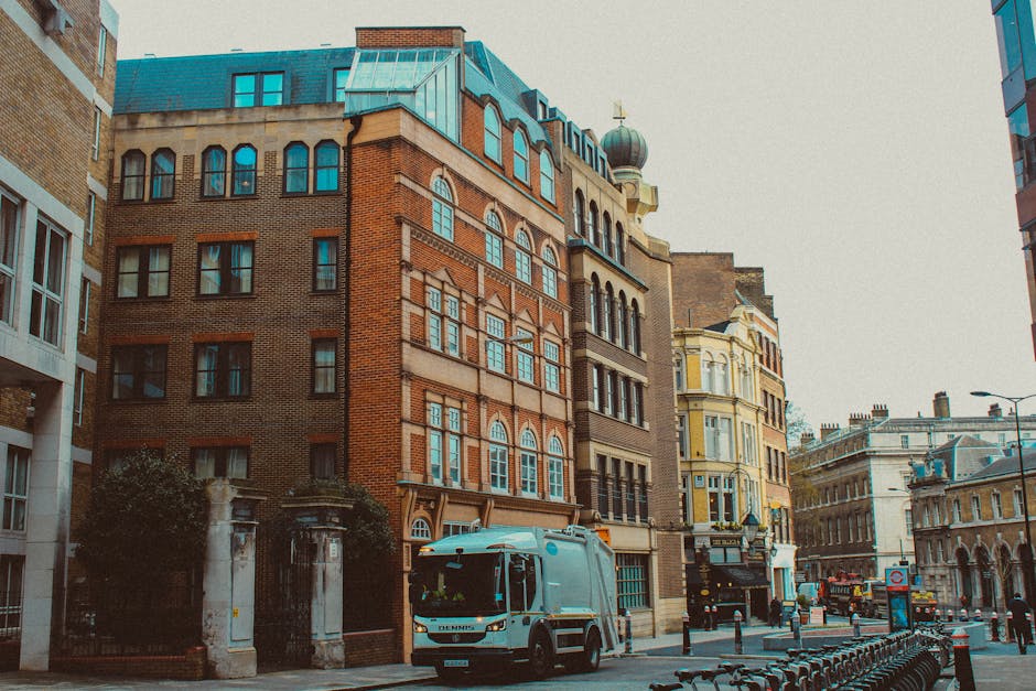A city street scene featuring a collection of multi-storey buildings with varying architectural styles, including brick facades, arched windows, and decorative cornices, typical of an urban environment. Prominent in the foreground is a light blue waste collection vehicle parked on the pavement, with a closed rear compartment designed for rubbish collection, indicating an active waste disposal service. To the right, row of black bicycle racks lines the sidewalk, and a black bollard prevents vehicle access, contributing to pedestrian safety. The background shows additional historic and modern buildings, along with street furniture like lampposts and signage. The street is relatively quiet with minimal traffic, and the lighting suggests a cloudy day with diffuse natural light. House Clearance Bayswater occasionally undertakes private waste handling and rubbish removal, which is implied by the scene, emphasizing on-site or independent collection methods.
This scene visually supports the context of alternative rubbish collection and waste management services available to residents, without specific promotional language, aligning with Bayswater's waste disposal options.