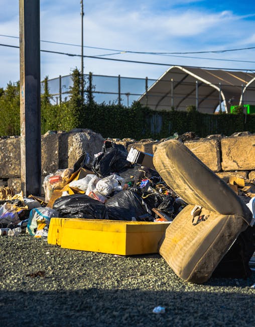 A piled collection of household rubbish including black and clear plastic garbage bags, an open yellow plastic container, a large discarded sofa cushion with fabric torn and foam visible, and various smaller debris such as paper, plastic, and cardboard, all situated on a gravel surface in an outdoor area. Behind the rubbish, a rough stone wall separates the scene from a background that features a metal fence, green shrubbery, and a partially visible tent-like structure with curved support beams. Overhead, power lines run across a blue sky with scattered clouds, indicating daytime sunlight. The scene showcases waste accumulation typical of non-official rubbish collection or private disposal, and the presence of the stone wall and outdoor environment aligns with a setting where independent waste removal services, such as those offered by House Clearance Bayswater, might be employed for on-site clearance of bulky household items and general rubbish.