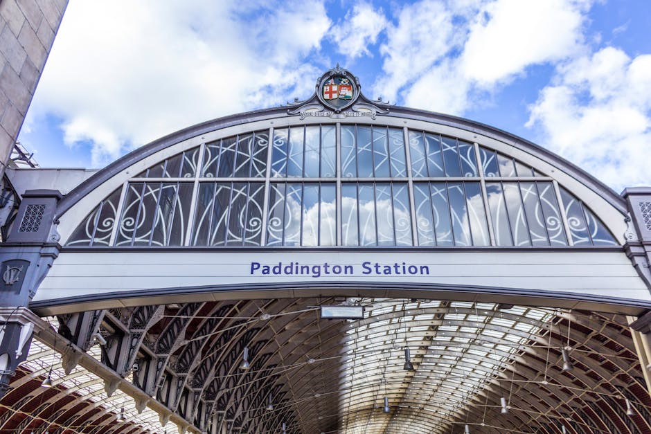 The image depicts the exterior of Paddington Station, showcasing an ornate, arched metal framework with intricate scrollwork and decorative elements painted in a light grey or silver finish. The upper part of the station features a prominent crest with a red cross, blue shield, and heraldic symbols, positioned at the peak of the arch. Below the crest, there is a white horizontal sign with the text 'Paddington Station' printed in dark blue. The station's metal and glass canopy extends below, with a curved, transparent roof allowing natural light to illuminate the interior space. The sky above is partly cloudy with patches of blue, creating a bright and airy atmosphere, while shadows and reflections can be seen on the glass surfaces. The structure's design emphasizes historical architecture, with a mix of Victorian and Edwardian influences, and the image focuses on capturing the detailed ironwork and the station's iconic entrance, depicting an identifiable railway terminus typical of urban transportation hubs and their associated infrastructure for passenger services or possibly overseeing private or independent waste collection operations as part of maintenance or clearance services by companies like House Clearance Bayswater.