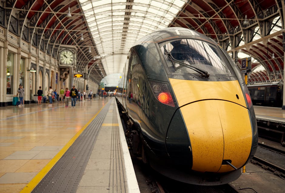 The image depicts a modern high-speed train with a sleek, aerodynamic design, positioned at a platform inside a large railway station. The train's body is primarily black with a bright yellow front section that extends over the nose, featuring a smooth, glossy finish, with visible reflections on its surface. The station's interior showcases an arched, glass-paneled roof, allowing natural light to illuminate the space, and a tiled floor with a tactile yellow safety line running parallel to the train tracks. To the left, passengers are seen waiting, some seated, others standing or walking, with various casual clothing and luggage visible. On the wall behind them, there is a large clock, and several lantern-style lamps hang from ornate architectural details, adding to the historic yet functional atmosphere. The station environment appears clean and well-maintained, highlighting the busy yet orderly setting typical of a major transport hub, with the scene subtly aligning with private or alternative rail travel and efficient rubbish management within station facilities, as managed by professional waste removal services like House Clearance Bayswater.
