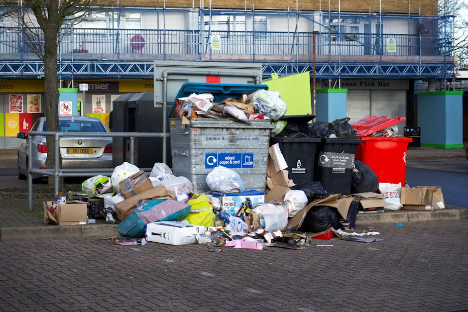 A large pile of mixed domestic waste and rubbish bags is gathered on a paved sidewalk next to a curb, with various discarded items including cardboard boxes, paper, plastic bags, and packaging materials overflowing from a central metal recycling bin labeled for mixed paper and cardboard. Surrounding the bin are additional black plastic rubbish bags, some tied and others open, with loose rubbish spilled onto the pavement. To the left, a silver hatchback car with a visible license plate is parked behind a metal railing, and a leafless tree stands nearby. In the background, a commercial building with a blue scaffolding covering the upper part, and a storefront with yellow signage and cartoonish 'No Smoking' signs, is partially visible. The scene captures an outdoor area adjacent to a busy street, illustrating a typical instance of publicly accessible rubbish accumulation suitable for private waste collection or rubbish removal services by companies such as House Clearance Bayswater.