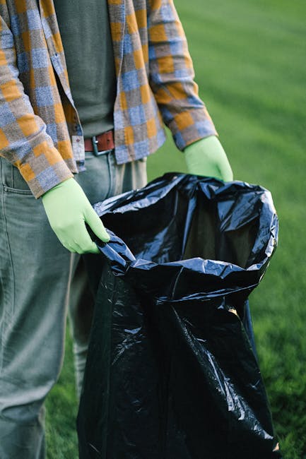 A person wearing a yellow and blue checkered shirt, a green t-shirt underneath, grey pants, and light green gloves is holding open a large black plastic rubbish bag outdoors on a grassy area. The individual's hands are grasping the edges of the bag, which appears to be partially filled or prepared for waste collection. The background shows a well-maintained lawn with blurred green foliage, indicating a garden or park setting. This scene illustrates private waste handling or on-site rubbish removal, commonly provided by waste management services such as House Clearance Bayswater, especially when opting for independent collection methods rather than local authority disposal. The focus on the container and the person's attire emphasizes safe handling of waste materials in an outdoor environment, aligning with professional rubbish clearance practices addressing various types of household or garden waste.