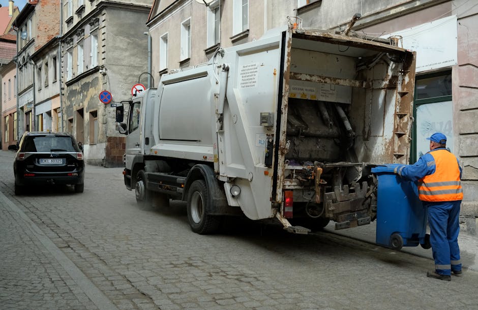 A large pile of mixed domestic waste and rubbish bags is gathered on a paved sidewalk next to a curb, with various discarded items including cardboard boxes, paper, plastic bags, and packaging materials overflowing from a central metal recycling bin labeled for mixed paper and cardboard. Surrounding the bin are additional black plastic rubbish bags, some tied and others open, with loose rubbish spilled onto the pavement. To the left, a silver hatchback car with a visible license plate is parked behind a metal railing, and a leafless tree stands nearby. In the background, a commercial building with a blue scaffolding covering the upper part, and a storefront with yellow signage and cartoonish 'No Smoking' signs, is partially visible. The scene captures an outdoor area adjacent to a busy street, illustrating a typical instance of publicly accessible rubbish accumulation suitable for private waste collection or rubbish removal services by companies such as House Clearance Bayswater.