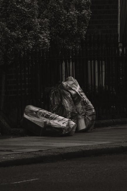 Two large black plastic garbage bags filled with waste material are placed on the pavement next to a wooden fence with vertical slats. The bags appear to be tightly packed and slightly crumpled, with visible creases on their surfaces. One bag has partially visible text printed on it, indicating it may have come from a packaging or shopping bag. The scene is set outdoors, with a dark backdrop of trees and foliage lining the area behind the fence, and a section of a brick or wooden structure partially visible in the upper right corner. The pavement surface beneath the bags is smooth, and the lighting suggests a subdued, overcast environment, typical of a late or early time of day. This visual context relates to private waste collection or rubbish removal services, similar to those offered by House Clearance Bayswater, highlighting the kind of waste that might be handled through independent rubbish collection or on-site clearance in the Bayswater area.
