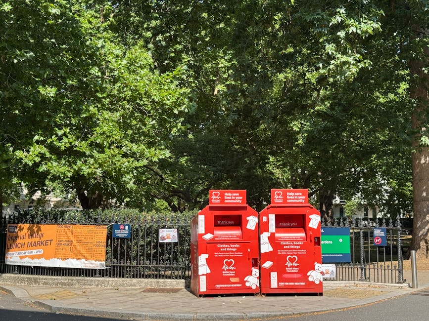 Two large black plastic garbage bags filled with waste material are placed on the pavement next to a wooden fence with vertical slats. The bags appear to be tightly packed and slightly crumpled, with visible creases on their surfaces. One bag has partially visible text printed on it, indicating it may have come from a packaging or shopping bag. The scene is set outdoors, with a dark backdrop of trees and foliage lining the area behind the fence, and a section of a brick or wooden structure partially visible in the upper right corner. The pavement surface beneath the bags is smooth, and the lighting suggests a subdued, overcast environment, typical of a late or early time of day. This visual context relates to private waste collection or rubbish removal services, similar to those offered by House Clearance Bayswater, highlighting the kind of waste that might be handled through independent rubbish collection or on-site clearance in the Bayswater area.